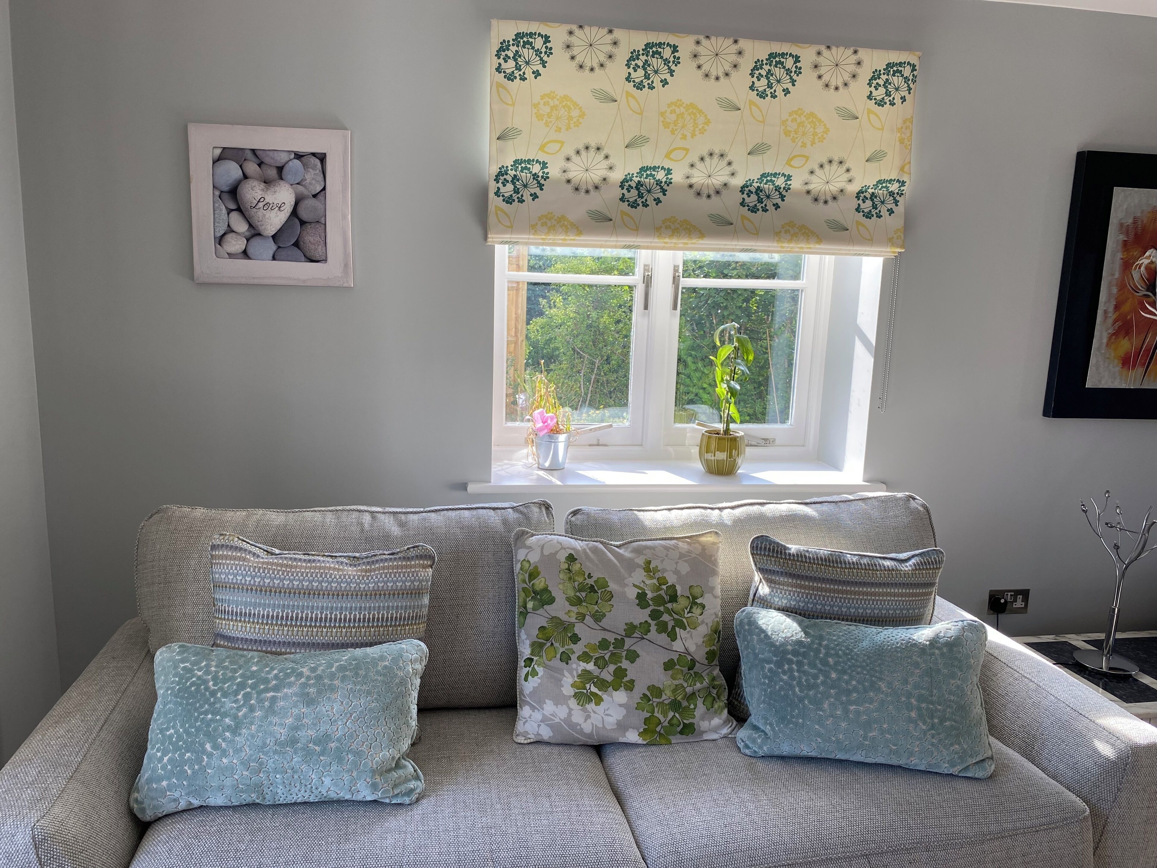 A living room with a grey sofa, patterned cushions, a framed picture of heart-shaped stones, a window with a floral valance, and a potted plant on the windowsill.