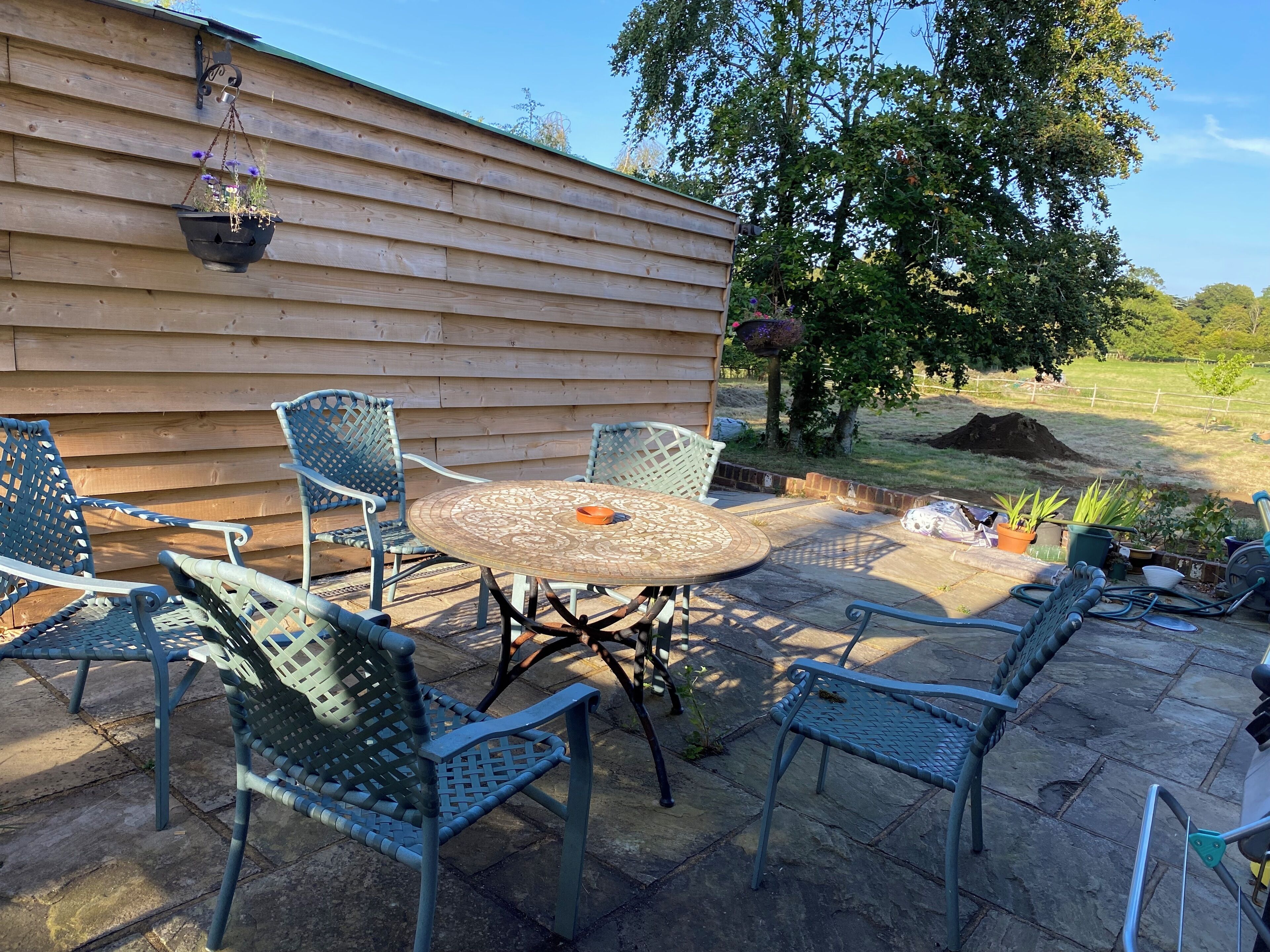A patio with a round table and four chairs, a hanging flower pot, and a wooden wall in the background.