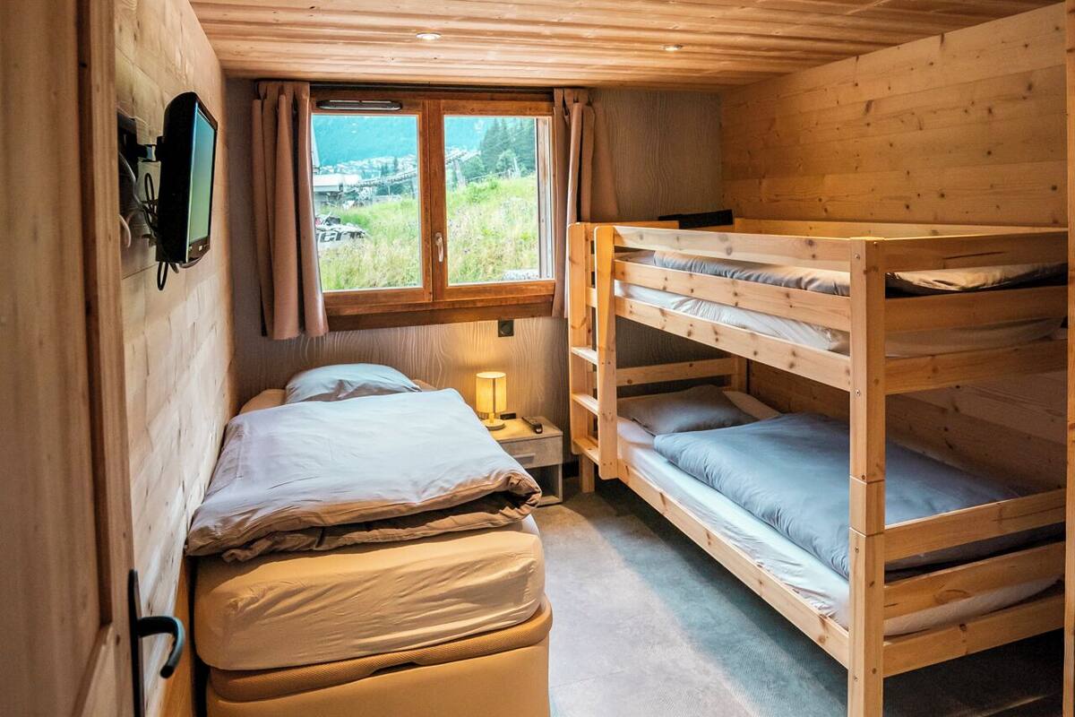 Ein Zimmer in einer Holzhütte mit Etagenbetten, einem Einzelbett, einem an der Wand befestigten Fernseher und einem Fenster mit Blick auf Grünflächen.