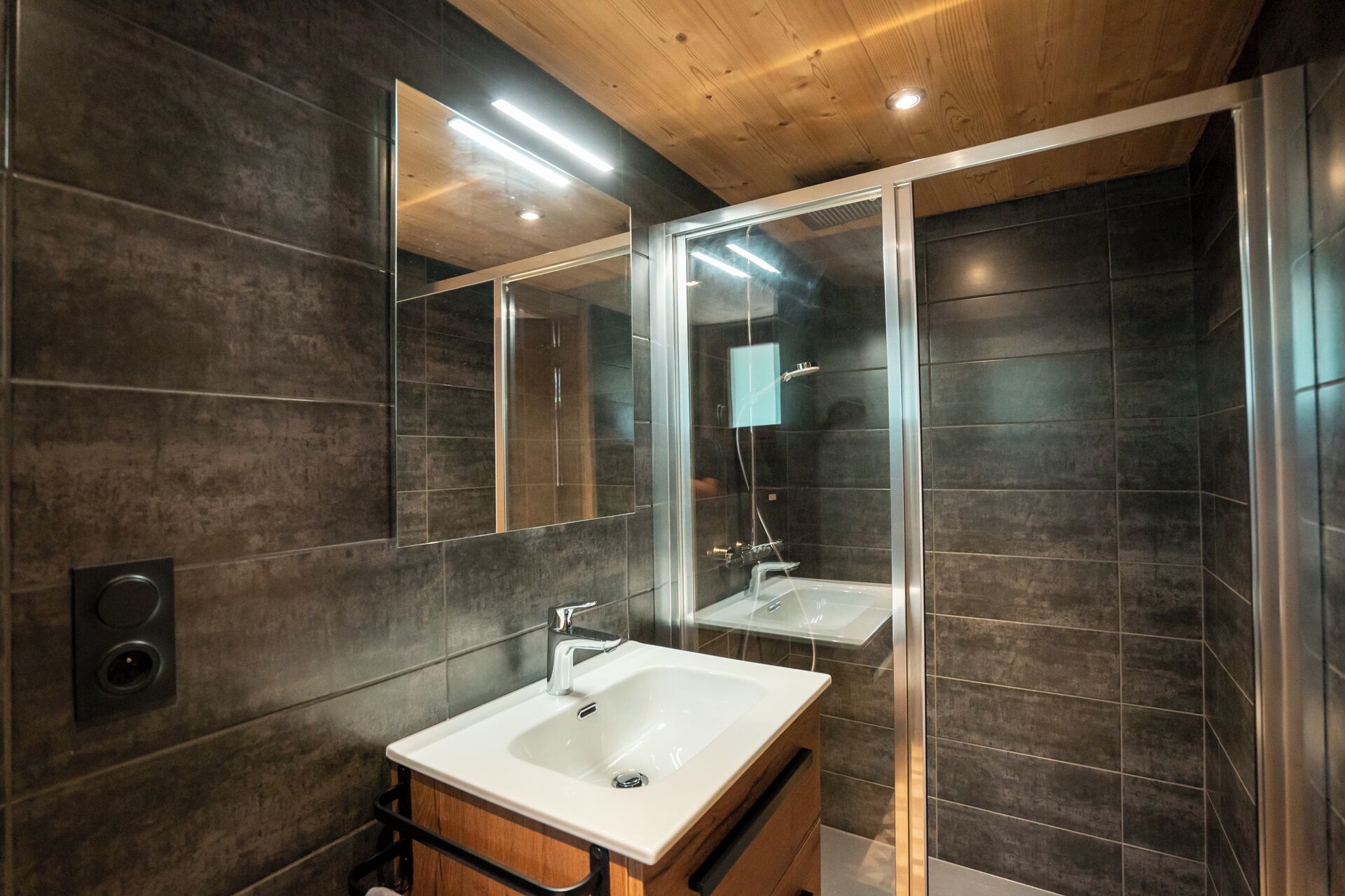 Ein modernes Badezimmer mit dunkel gefliesten Wänden, einer Holzbalkendecke, einem großen Spiegel, einem weißen Waschbecken und einer Duschkabine aus Glas.
