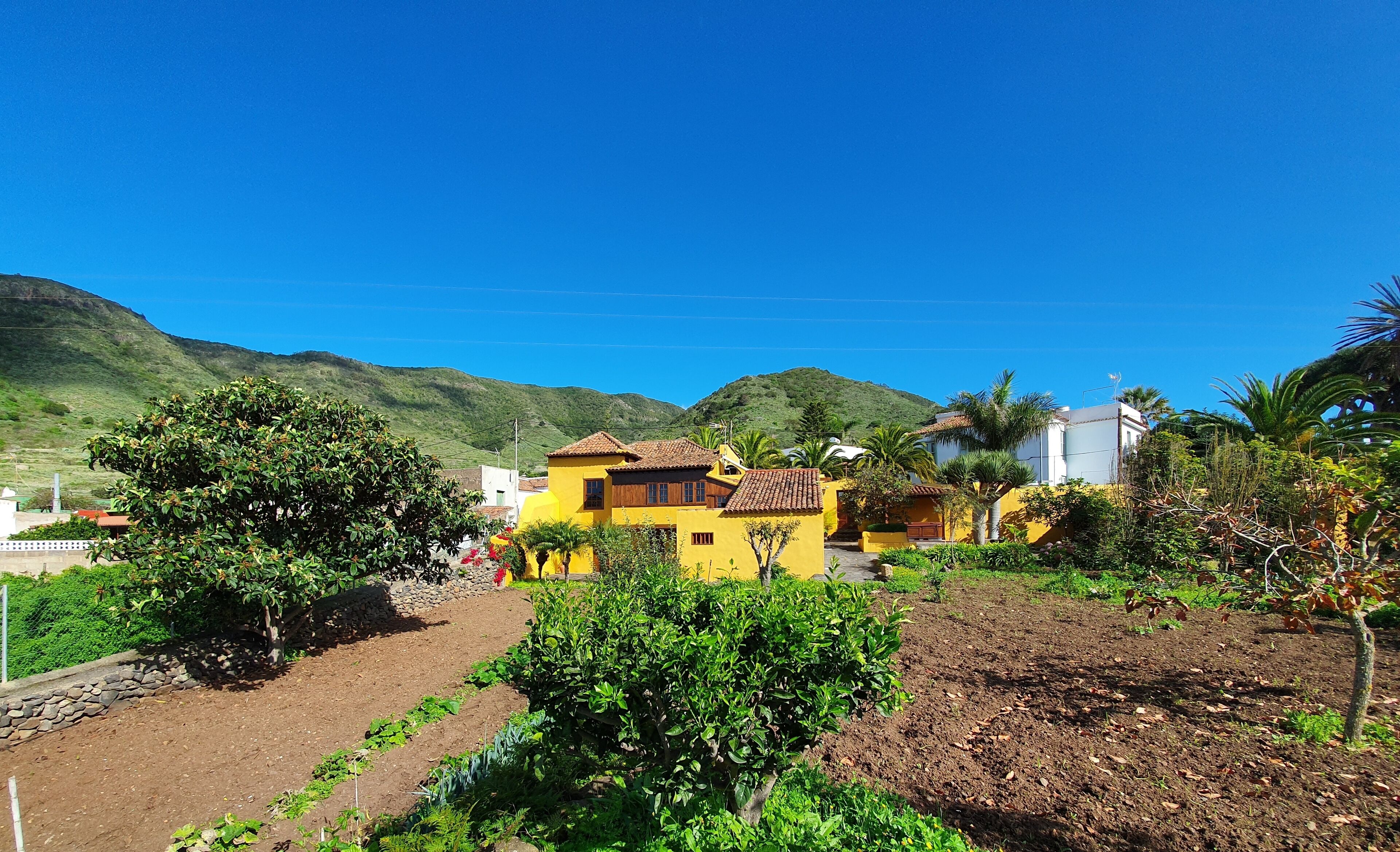 Un edificio amarillo con techo de tejas rojas, rodeado de vegetación y un paisaje montañoso.