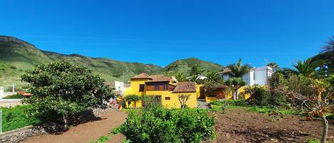Un edificio amarillo con techo de tejas rojas, rodeado de vegetación y un paisaje montañoso.