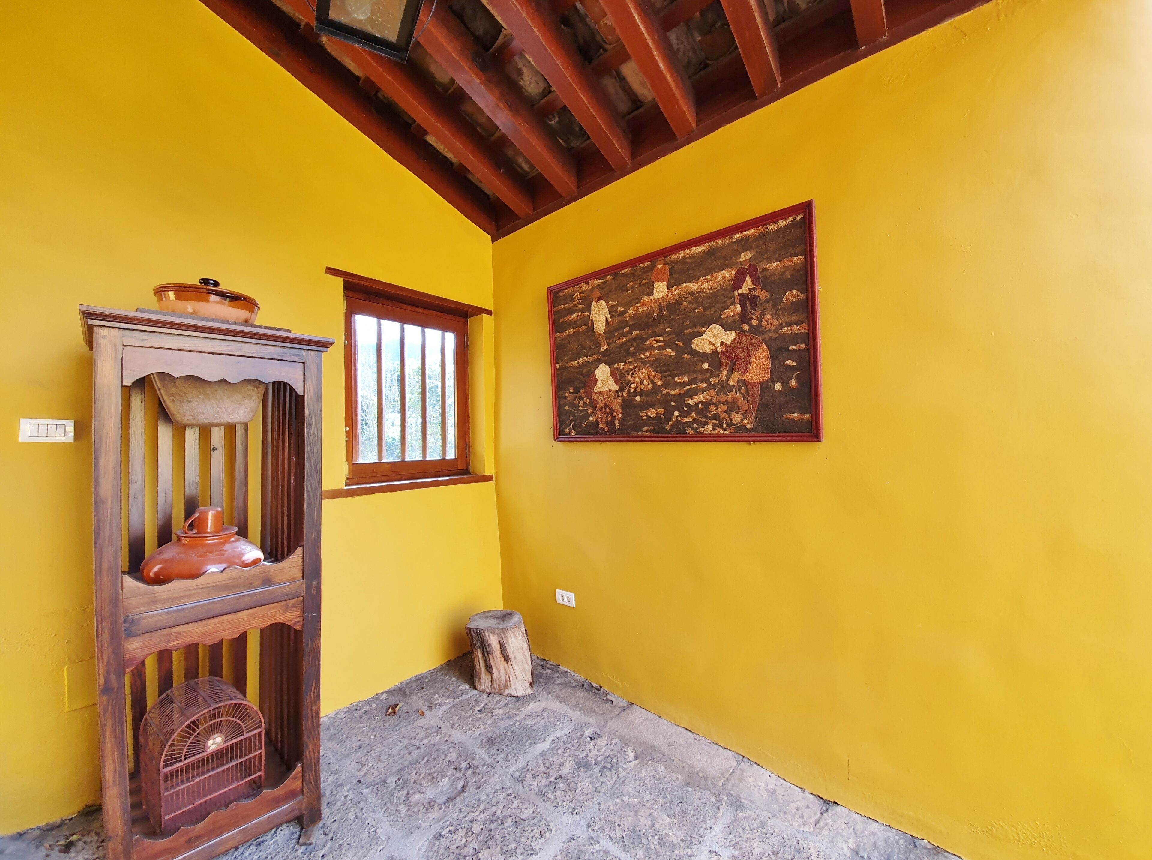 Una habitación con paredes amarillas, techo de madera, una ventana, un cuadro, un armario de madera y un taburete pequeño.