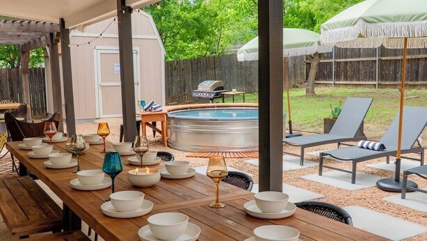 Un área de comedor al aire libre con una larga mesa de madera dispuesta para una comida, rodeada de sillones y sombrillas.