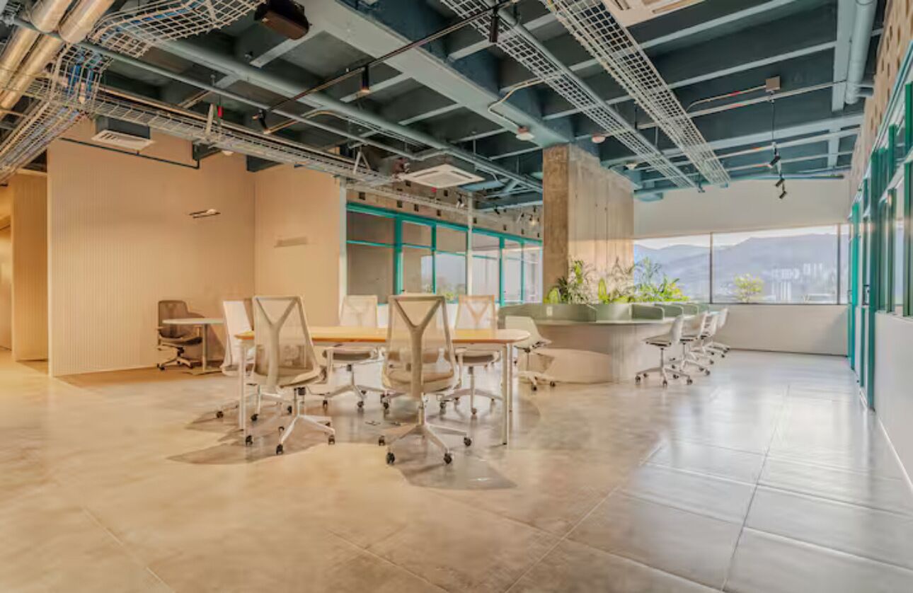 A spacious office with a high ceiling, exposed ductwork, and large windows offering a view of the city.