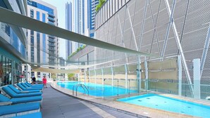 A rooftop pool area with blue lounge chairs, a swimming pool, and a modern building with a geometric facade in the background. - Elegant 2-bed Retreat Stunning Downtown Views (Dubai)