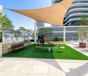 A rooftop garden with a children's play area, including a slide and swings, surrounded by a fence and potted plants.