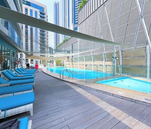 A rooftop pool area with blue lounge chairs, a swimming pool, and a modern building with a geometric facade in the background. - Elegant 2-bed Retreat Stunning Downtown Views (Dubai)
