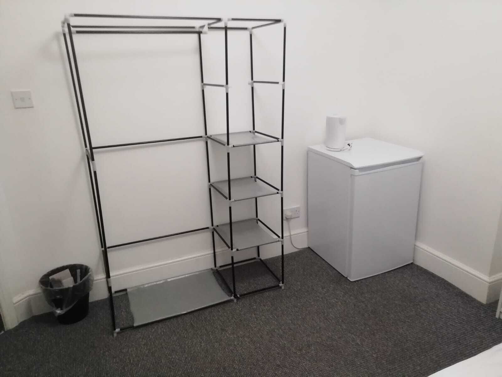 A corner of a room with a metal shelving unit, a small white washing machine, and a trash bin.