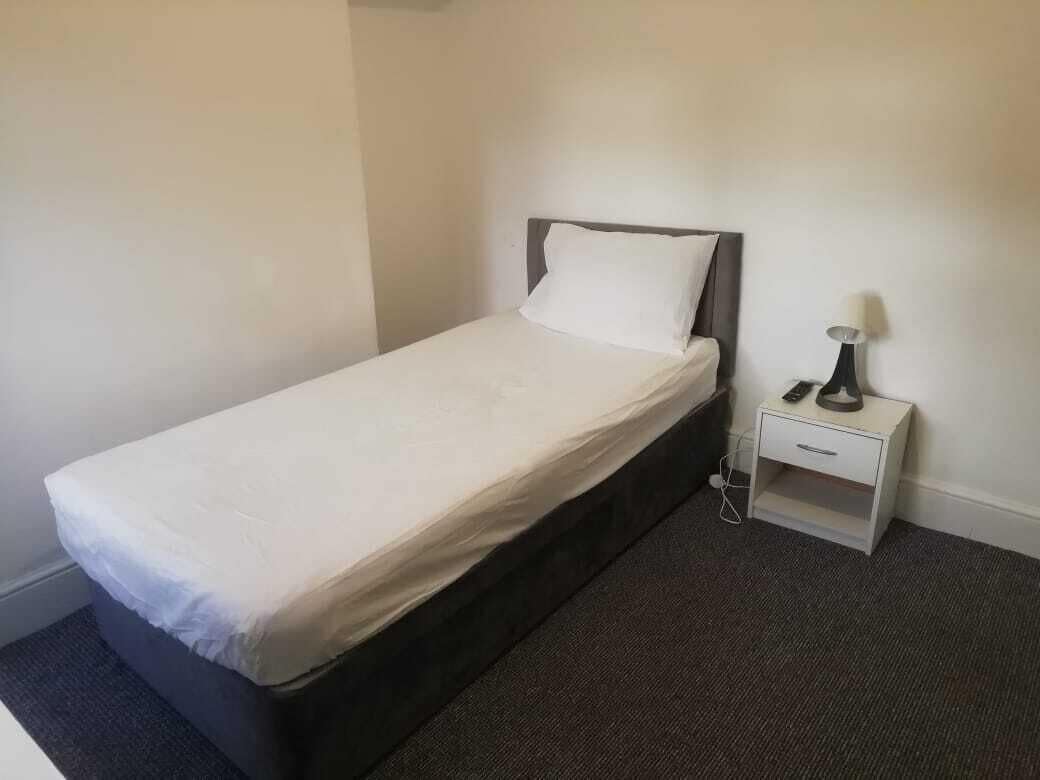 A single bed with a white mattress and pillow, a bedside table with a lamp, and a carpeted floor.