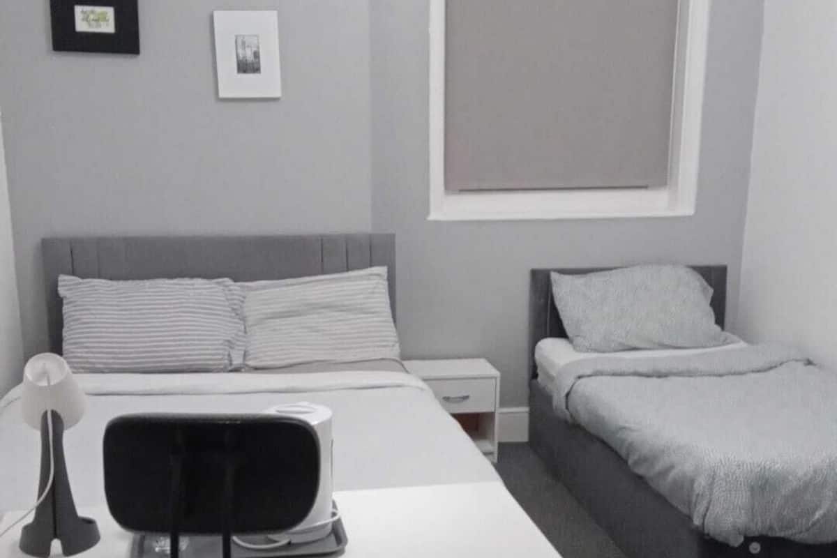 Una habitación pequeña y moderna con una cama, un escritorio y una silla. Hay dos cuadros enmarcados en la pared, una ventana y una puerta blanca.