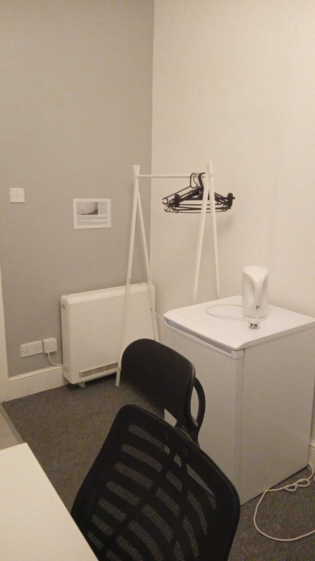 A small office space with a white desk, a black office chair, a white radiator, and a white electrical outlet on the wall.