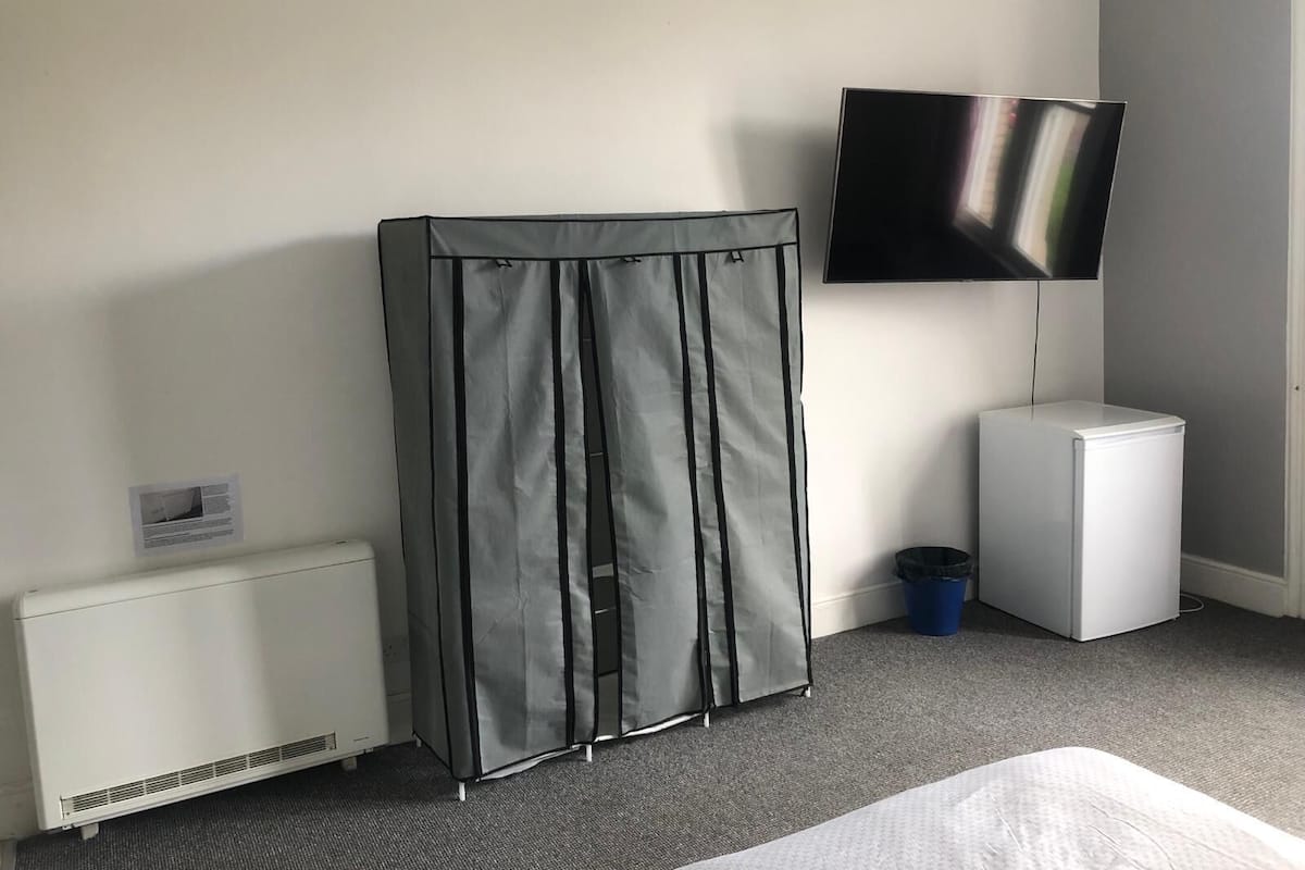Una habitación de hotel con cama, televisor sobre un soporte, calentador, cubo de basura y unidad de almacenamiento.