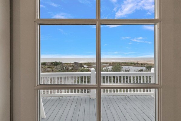 Vista a través de una ventana con marco blanco, que da a una terraza con barandilla blanca y un cielo azul despejado.
