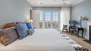 A bedroom with a large bed, a ceiling fan, a desk with a chair, and a view of the outside through a window.