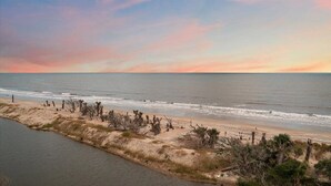 A coastal landscape with a beach, sparse vegetation, and a calm sea under a sunset sky.