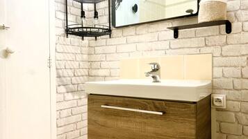 Un baño con pared de ladrillo blanco, un lavamanos blanco con gabinete de madera debajo, un estante metálico negro con una toalla y una botella, y un espejo sobre el lavamanos.