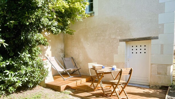 Een terras met twee lounge stoelen en een tafel, omringd door een tuin en een gebouw met een structuurmuur en een deur.