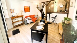 Een moderne woonkamer met een bank, eettafel en stoelen. Een trap leidt naar een keuken met een bakstenen muur en een raam.