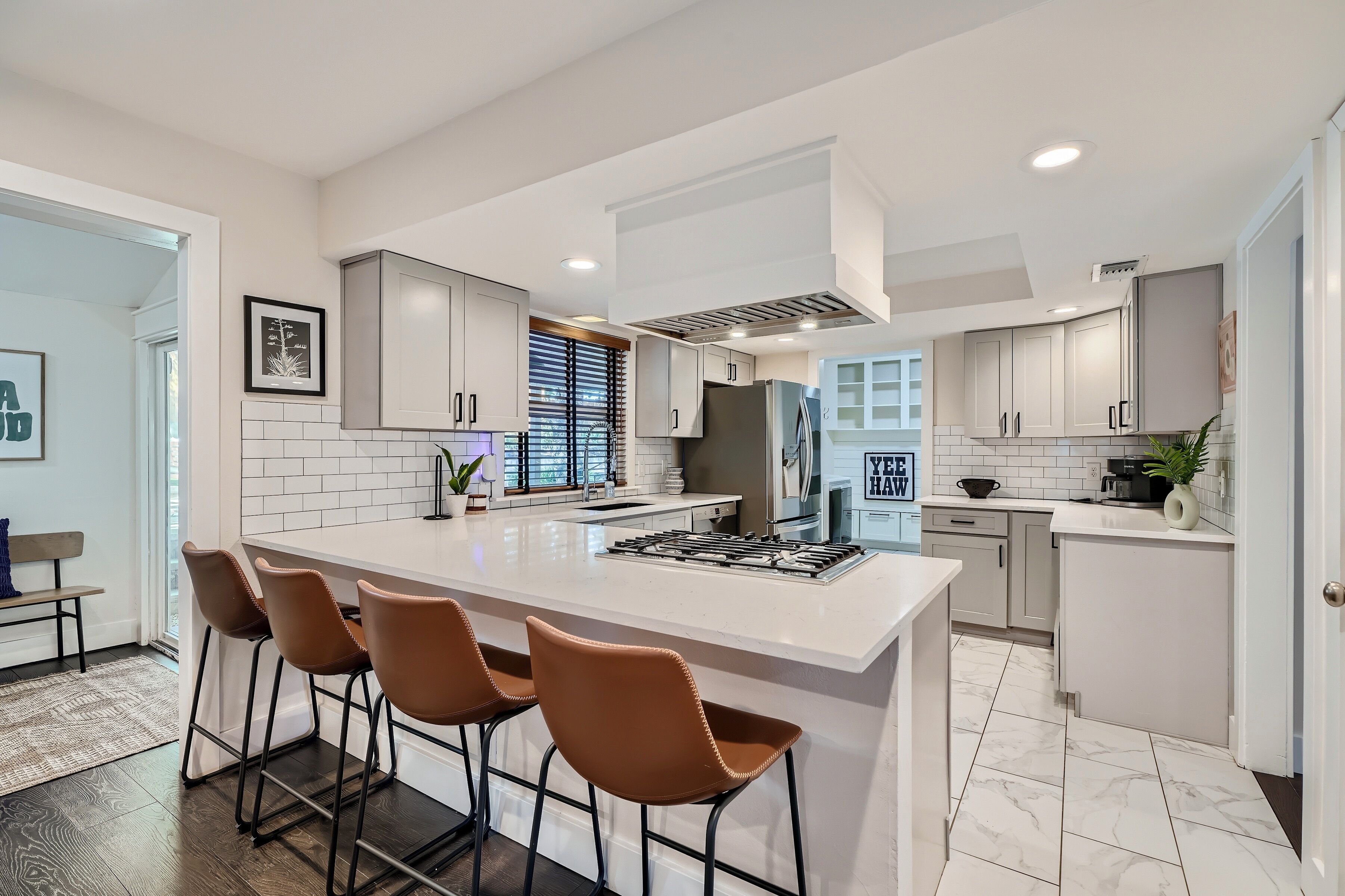 A modern kitchen with a central island, bar stools, white cabinetry, and a large window with blinds.
