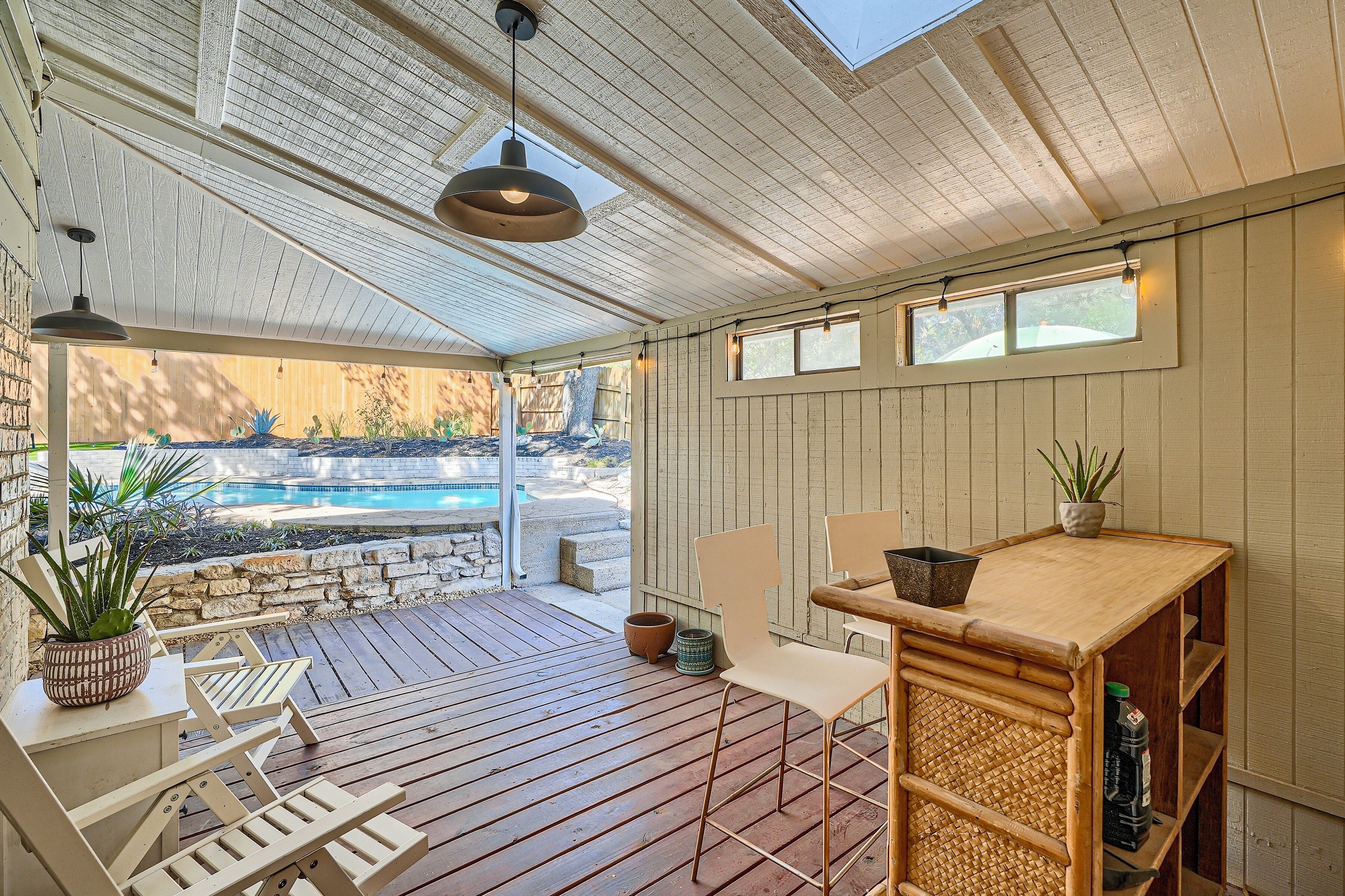A covered outdoor area with a pool, wooden deck, seating, and a table with a plant.