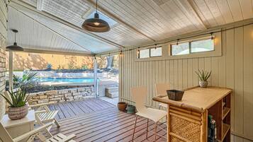 A covered outdoor area with a pool, wooden deck, seating, and a table with a plant.