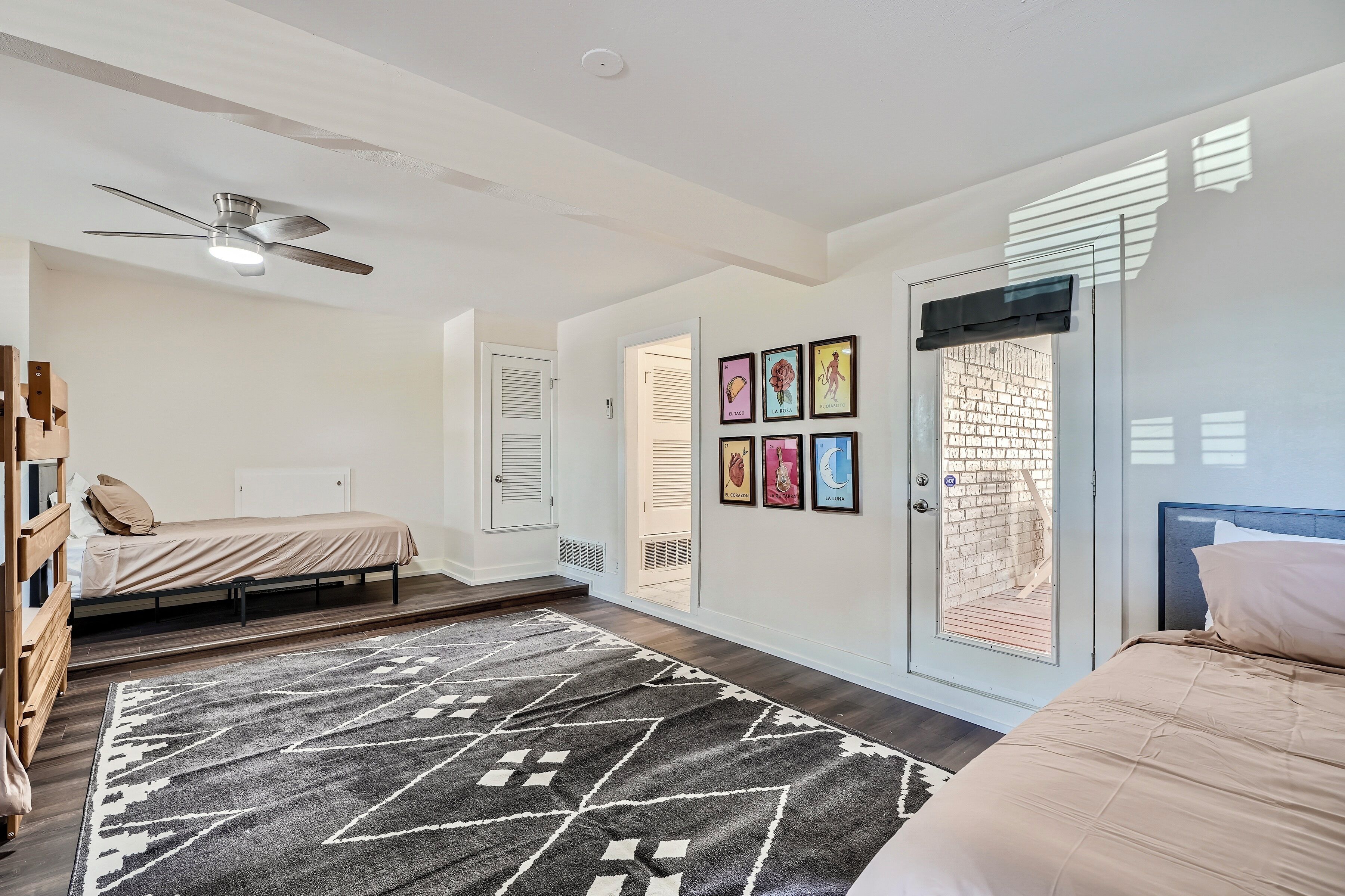 A bedroom with a bed, a ceiling fan, a door with a window, and a wall with framed pictures.