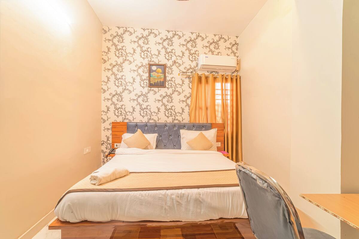 Uma cama bem arrumada com cabeceira de madeira, uma pequena mesa de cabeceira e uma cadeira com estrutura de metal.