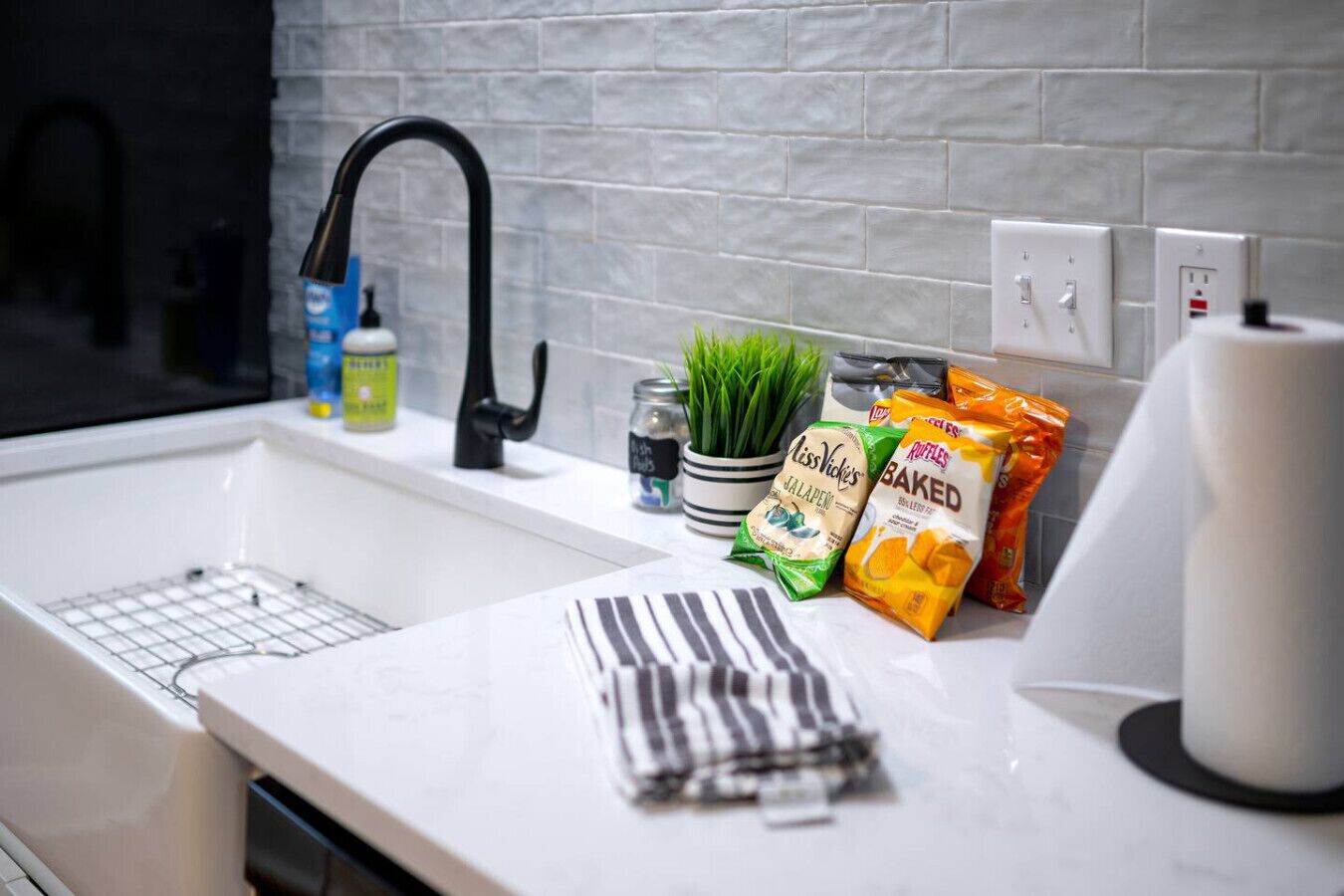 A kitchen with a white countertop, a black faucet, a white sink, and a white tiled backsplash. There are bags of snacks and a jar of green herbs on the counter.
