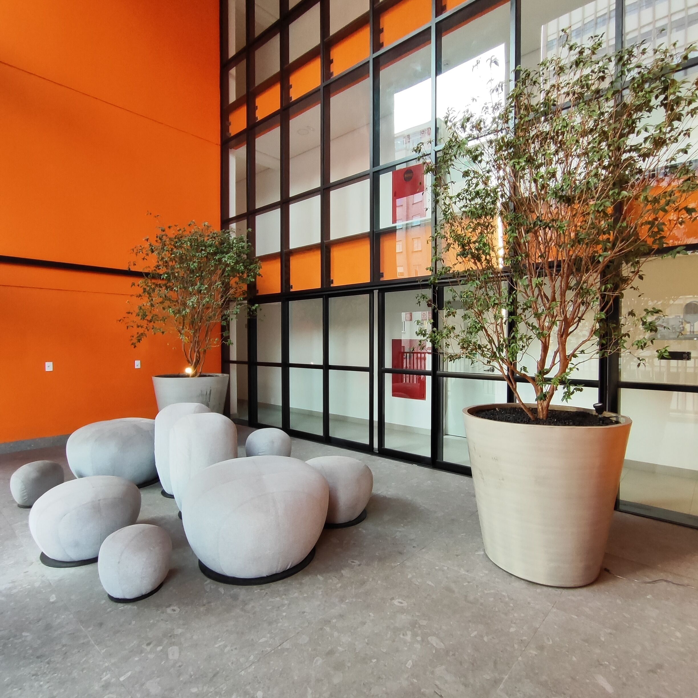 Um interior moderno com grandes janelas, pufes cinzas e plantas em vasos.