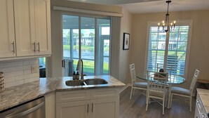 A modern kitchen with white cabinetry, a stainless steel dishwasher, and a sink. There is a dining area with a glass table and white chairs. - Gated community condo with private island minutes to Fort Myers Beach or Sanibel (Fort Myers)