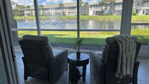 A view from a room with large windows overlooking a water feature and a building complex. There are two wicker chairs and a small table with a plant. - Gated community condo with private island minutes to Fort Myers Beach or Sanibel (Fort Myers)