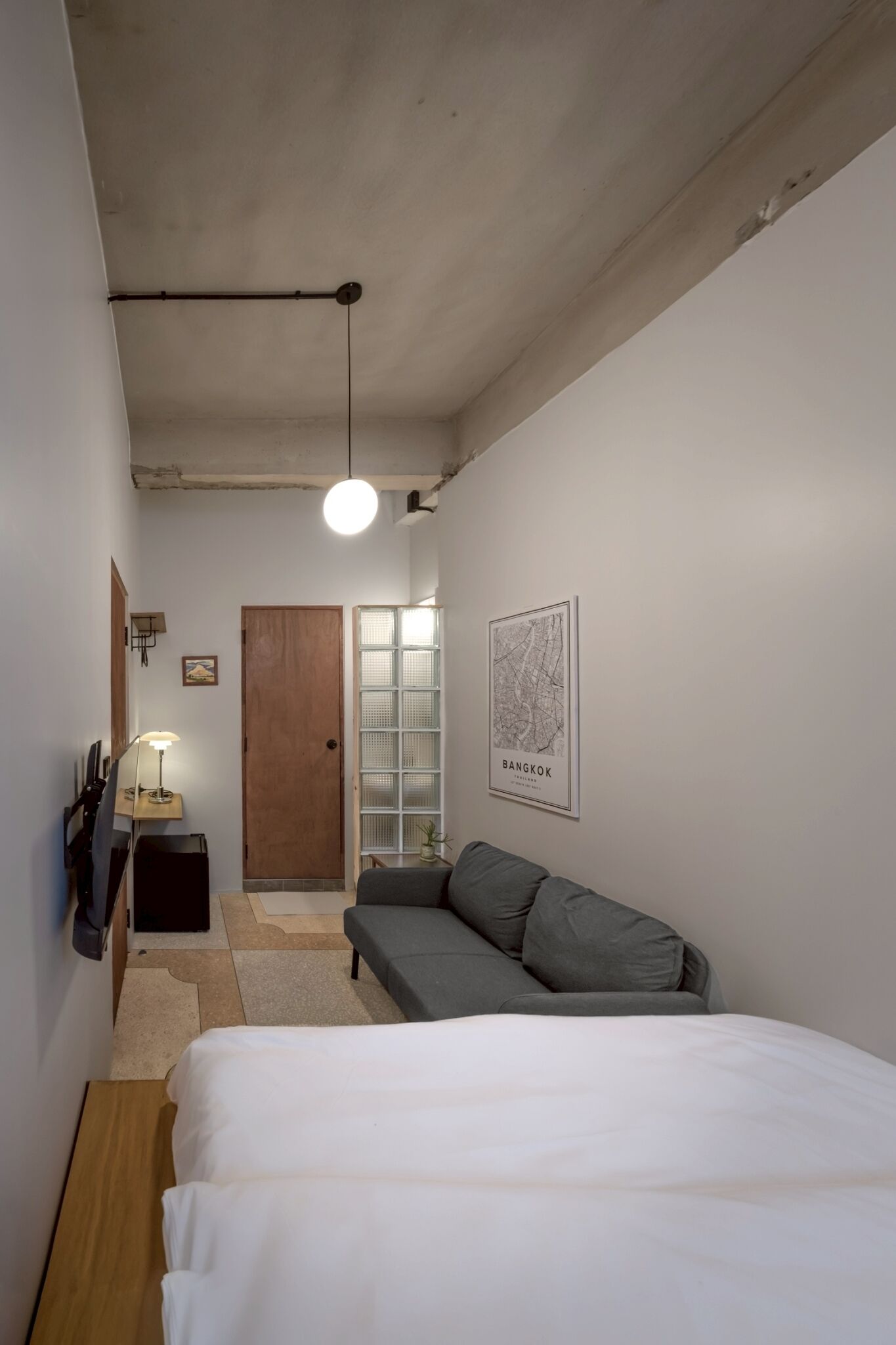 Un estrecho pasillo que conduce a un dormitorio con una cama, un sofá gris, una puerta de madera y un mapa de Bangkok en la pared.
