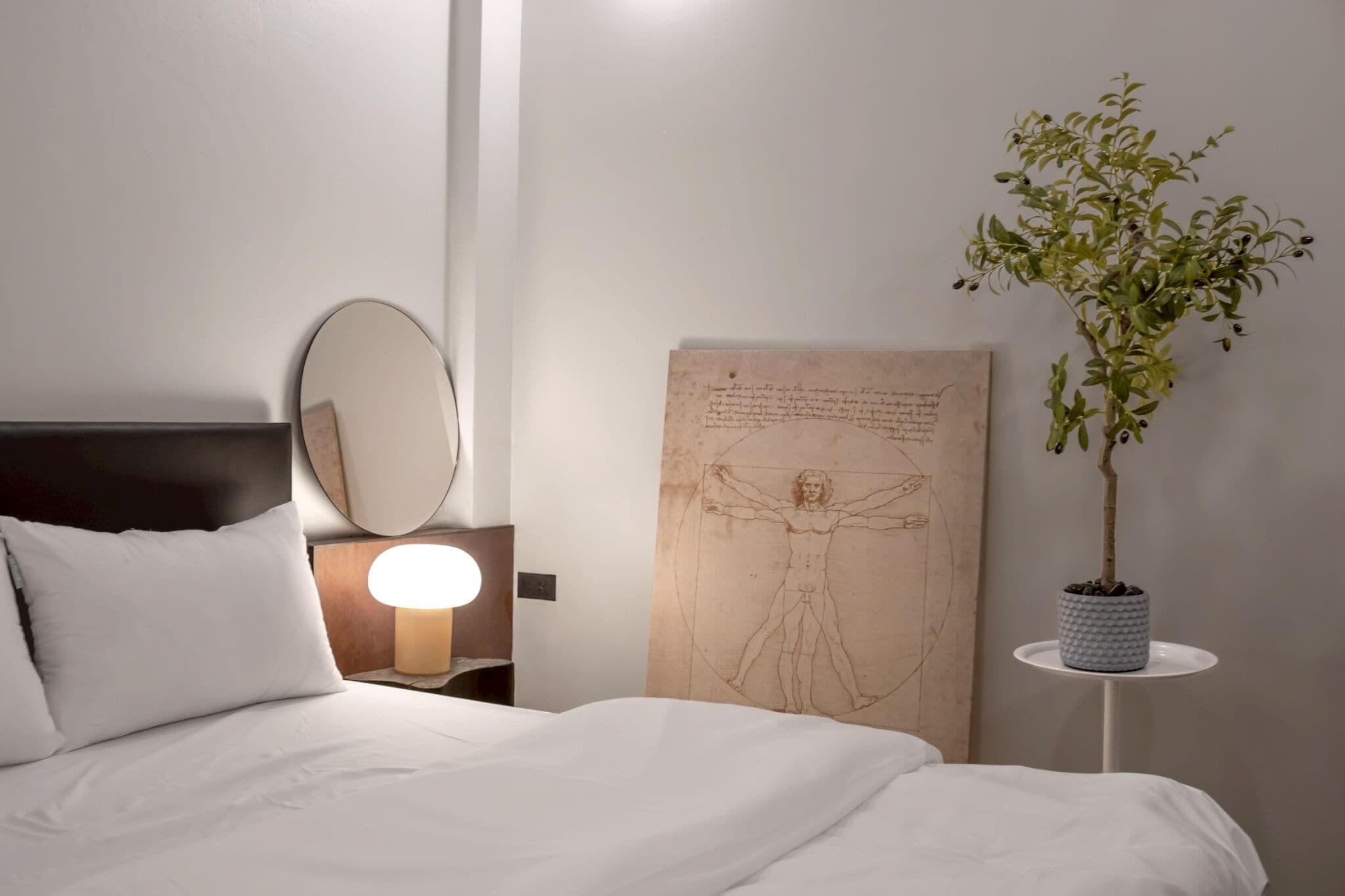 A bedroom with a bed, a bedside lamp, a wall-mounted mirror, and a framed sketch on the wall.