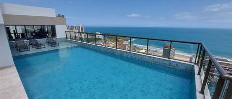A rooftop infinity pool with a city view, lounge chairs, and a clear sky.
