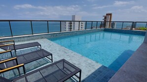 A rooftop infinity pool with glass railings, lounge chairs, and a view of the ocean and cityscape. - COBAR0129 - Edifício Barra Premium (Salvador)
