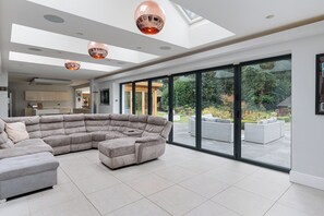 A spacious living room with a large sectional sofa, a dining area, and a view of the garden through large sliding glass doors. - Luxury Villa in Knutsford With Heated Indoor Pool & Hot Tub (Allostock)