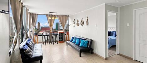 Sala de estar con sofá, una mesa de comedor con sillas, balcón con cortinas y vista panorámica de la ciudad.