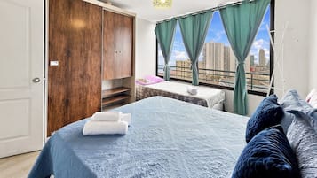 A bedroom with a large bed, a wooden wardrobe, a small seating area, and a balcony with a city view.
