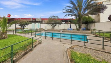 Una piscina con un paseo perimetral, palmeras y un edificio con techo rojo al fondo.