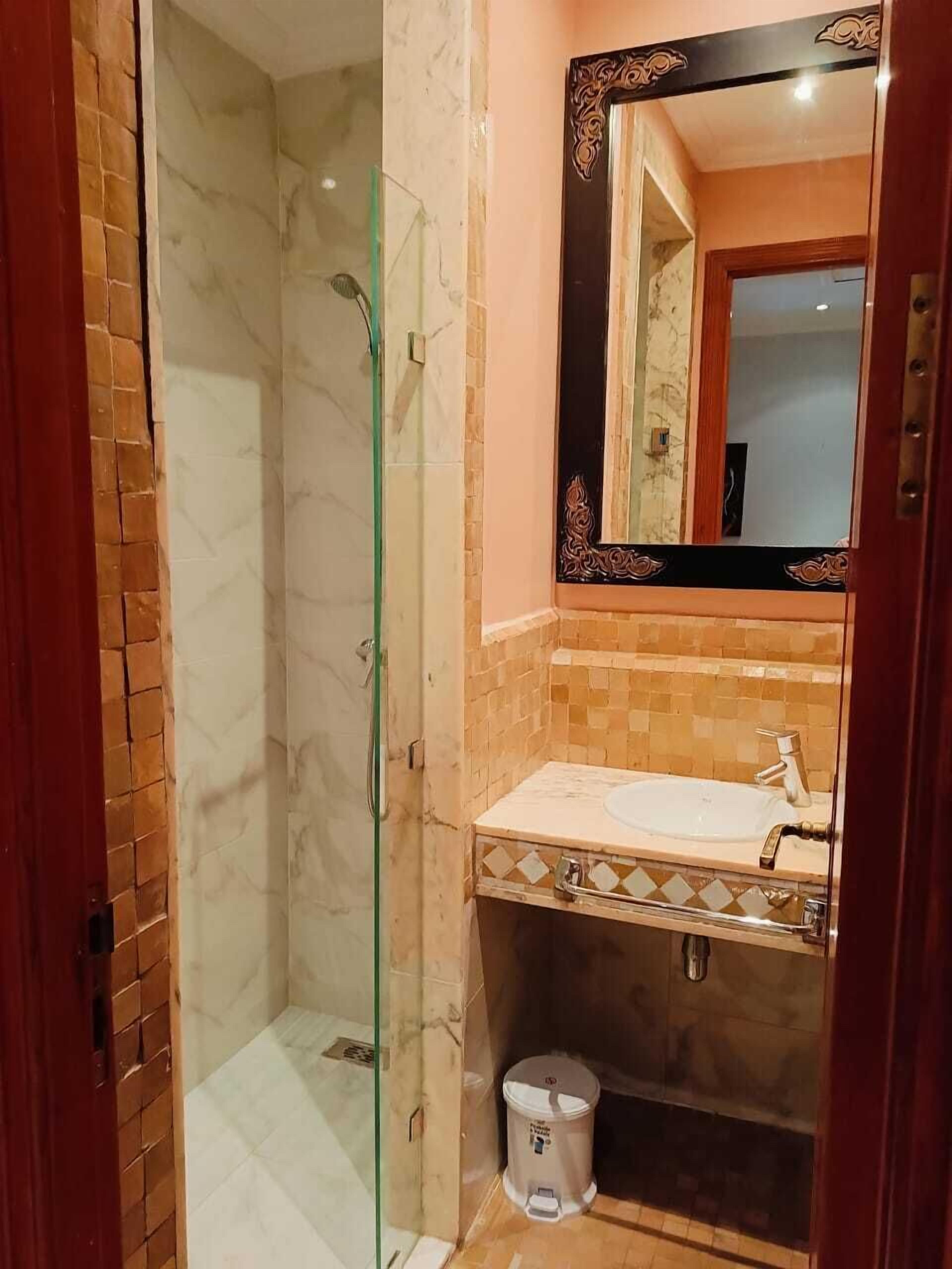 A bathroom with a glass-enclosed shower, a sink with a mirror, and a trash bin.