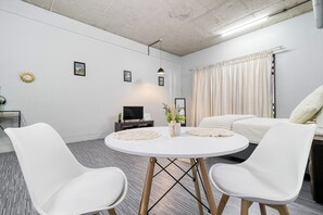 Une chambre d’hôtel moderne avec une table de salle à manger ronde, deux chaises blanches, un lit avec une literie blanche, un téléviseur et une plante en pot.