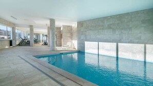 An indoor swimming pool with a tiled floor, a lounge chair, and exercise equipment in the background.