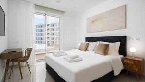 A hotel room with a bed, a desk, and a chair. There is a view of buildings outside the window.