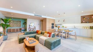 Un salon moderne avec un canapé d’angle, une table basse et un coin repas, la cuisine étant visible au fond.