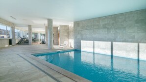 Une piscine intérieure dotée d’un sol et de murs carrelés, entourée de fenêtres et d’appareils de musculation.