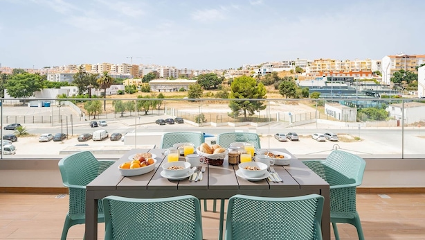 Un balcon avec une table dressée pour le petit-déjeuner, d’où l’on peut admirer un panorama urbain avec ses bâtiments et son parking.