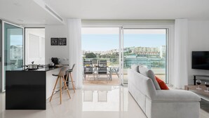 A modern living room with a sofa, a dining area with a table and chairs, a kitchen area with a black countertop, and a large sliding glass door leading to a balcony with a city view. - CourtView Modern Lagos Apartment (Lagos)
