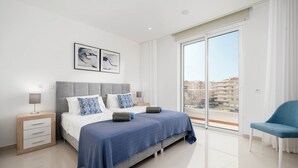 A modern bedroom with a large bed, a nightstand, a chair, and a view of buildings through a sliding glass door. - CourtView Modern Lagos Apartment (Lagos)