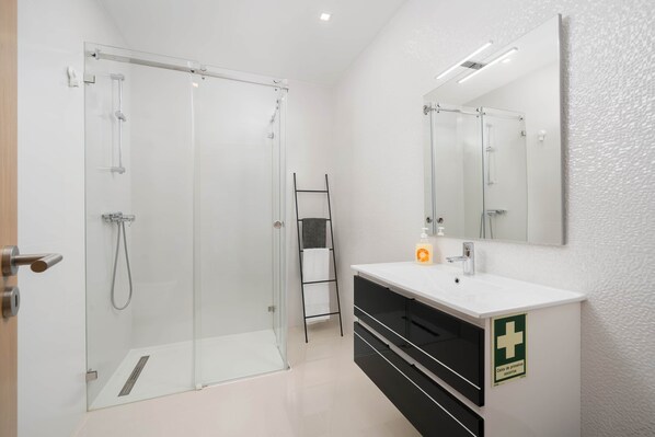 A modern bathroom with a glass shower enclosure, a white sink with a soap dispenser, and a first aid kit sign on the wall.